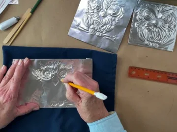 A pair of hands embossing the image of an owl into a sheet of aluminium foil.