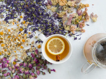 A cup with a slice of lemon and cinnomon quill, surrounded by dried rose petals, chamomile flowers and pea flowers.