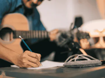 A man holding a guitar whilst writing in a notebook
