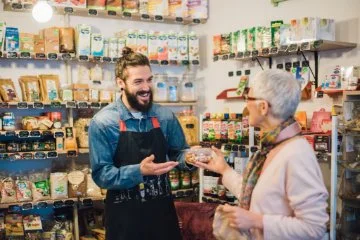 A man with a beard and a man-bun handing groceries to an older lady