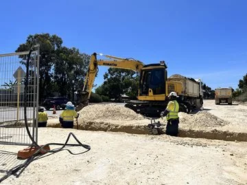 Three people stand in a trench and dig. They are wearing high-visibility clothing and hard hats. Behind them, a large excavator scoops up sand.