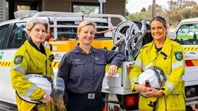 Three women stand in front of n emergency vehicle in fire protective gear