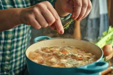 A person places some fresh herbs into a pot of hot soup.