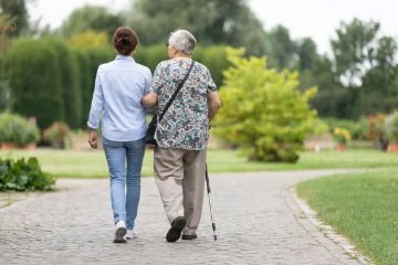 Two people walk side by side along a garden path, one using a walking stick, surrounded by greenery and landscaped plants.