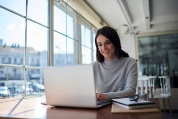 Person sitting at a wooden table near large sunlit windows, using a laptop, with a notebook and glasses placed beside the laptop in a bright, modern indoor workspace.