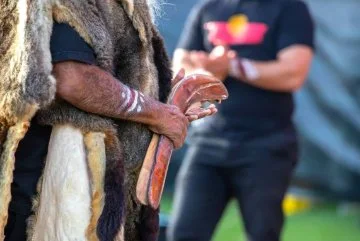 A person holds a wooden boomerang, with another figure blurred in the background during an activity outdoors.
