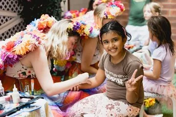 A young girl gets her arm painted by a woman dressed in bright colours.